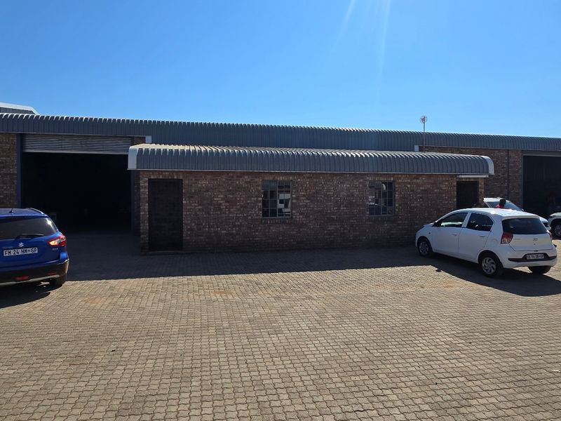 Front view of an industrial warehouse property featuring face brick construction, corrugated metal roofing, and multiple roller shutter doors with a paved yard area.
