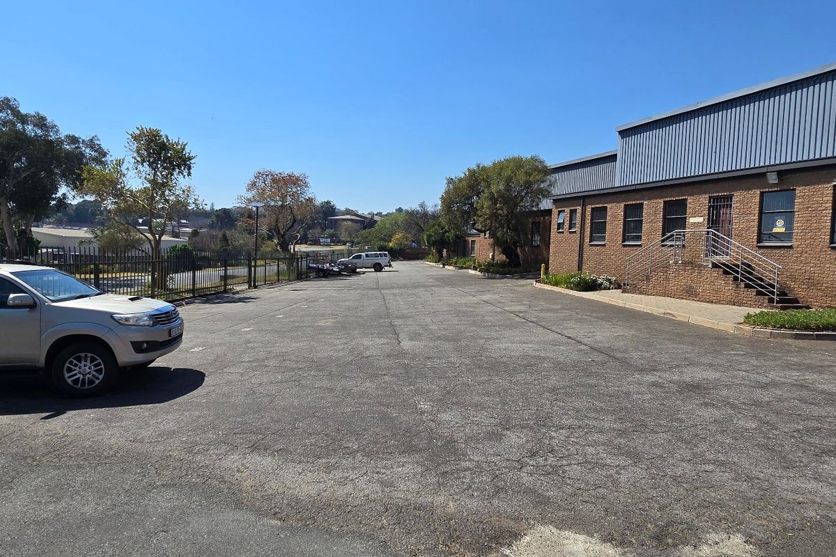 Exterior view of an industrial property featuring a brick facade, metal roofing, and a large asphalt parking area with security fencing.