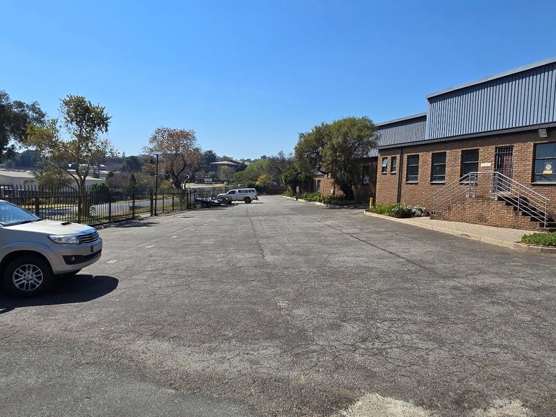 Exterior view of an industrial property featuring a brick facade, metal roofing, and a large asphalt parking area with security fencing.