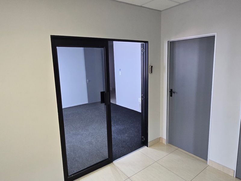 A clean, modern commercial hallway featuring tiled flooring, internal doors, and a glass sliding door leading into an office space.