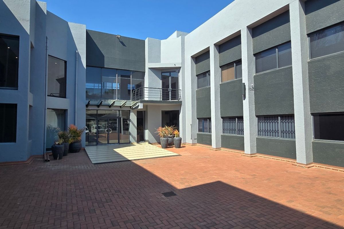 A modern commercial courtyard featuring paved flooring, a glass-fronted building facade, and secure metal window treatments.