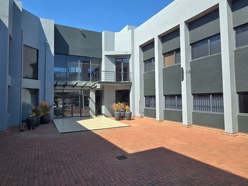 A modern commercial courtyard featuring paved flooring, a glass-fronted building facade, and secure metal window treatments.