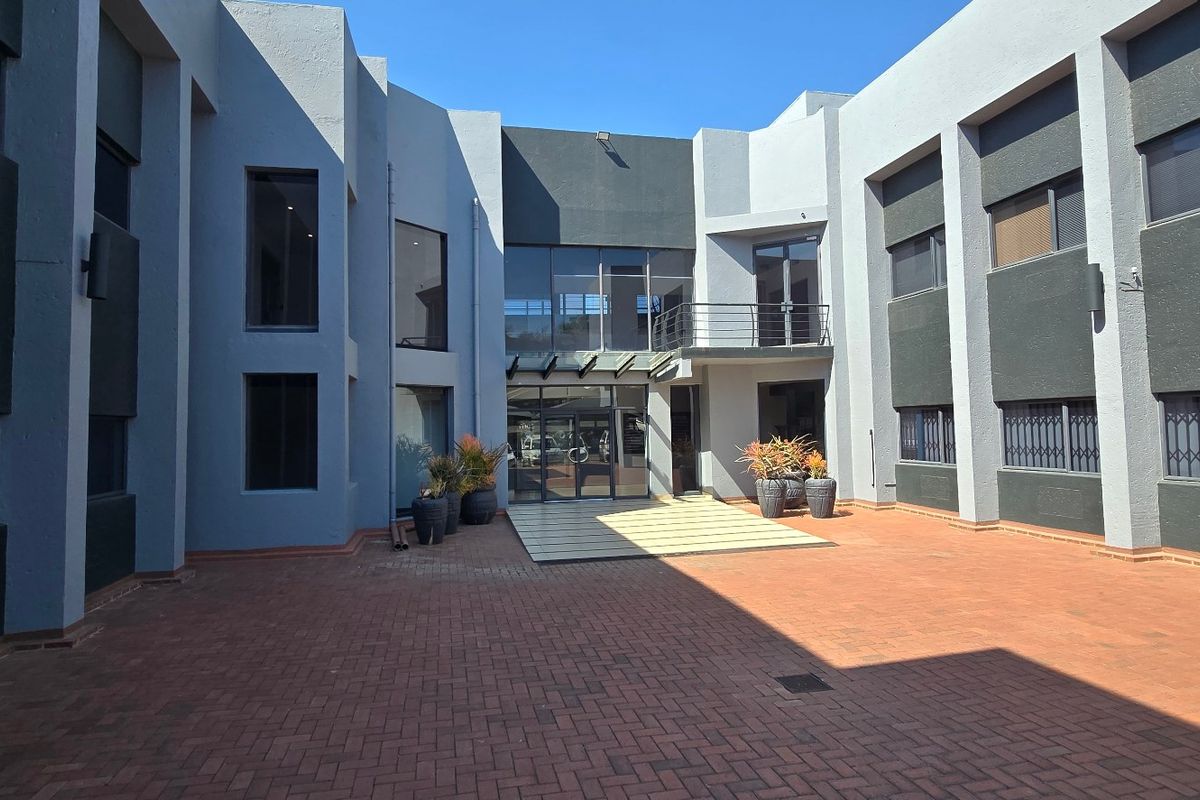 A spacious commercial courtyard featuring paved flooring, modern glass facade, and multi-level office building access with secure window treatments.