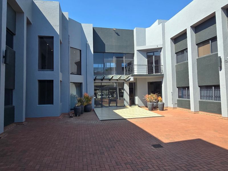 A spacious commercial courtyard featuring paved flooring, modern glass facade, and multi-level office building access with secure window treatments.