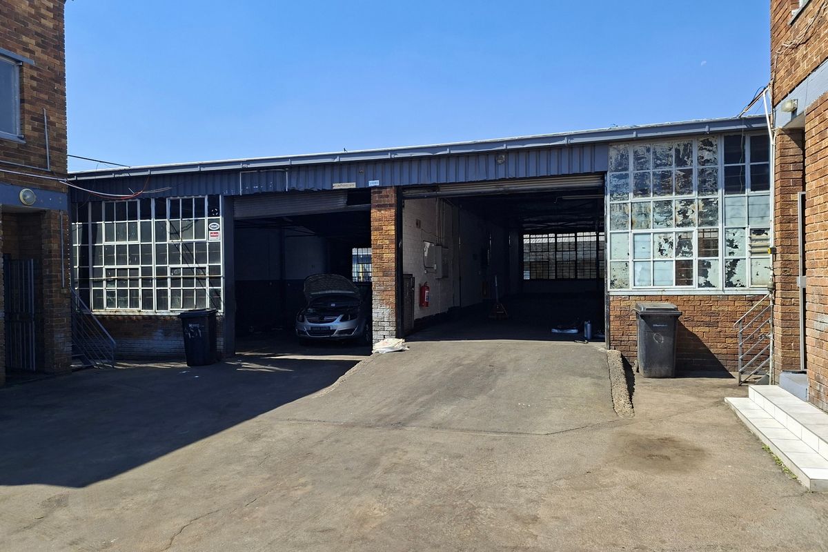 Industrial rear exterior view featuring large roller shutter access doors, steel-framed windows, and a concrete driveway area.