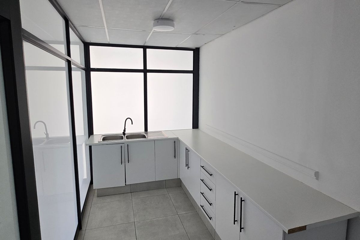 A clean, modern kitchenette featuring white built-in cabinetry, a stainless steel sink, tiled flooring, and a glass partition wall in a commercial office space.
