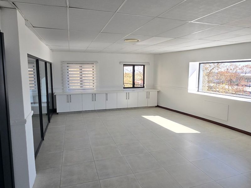 A bright, vacant commercial office space featuring tiled flooring, a suspended ceiling, built-in storage cabinetry, and large windows with roller blinds.