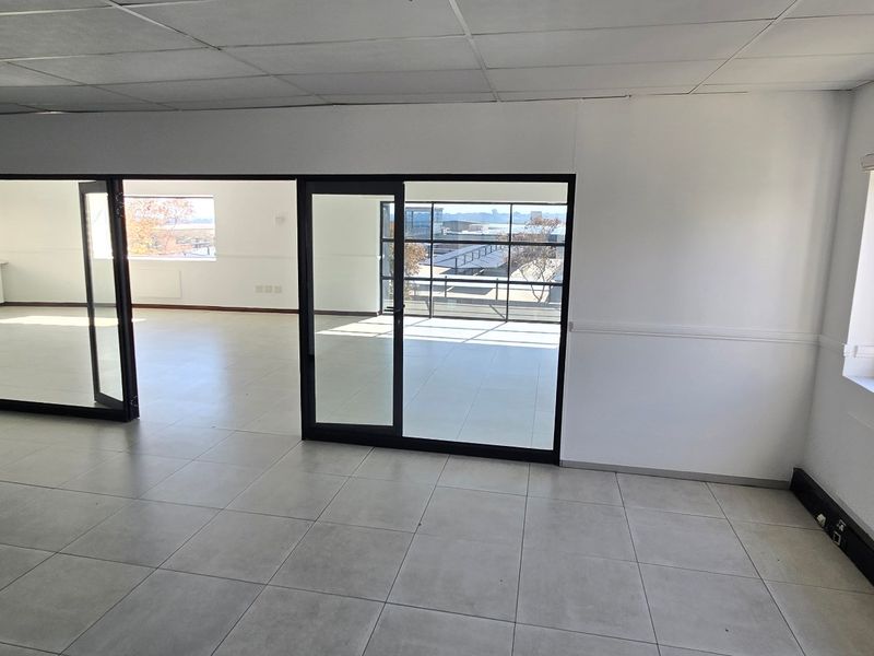 A bright, open-plan commercial office space featuring neutral tiled flooring, a suspended ceiling, and large sliding glass doors providing natural light and exterior views.