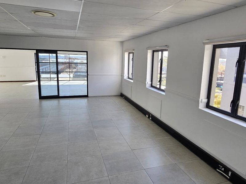 A bright, open-plan commercial office space featuring neutral tiled flooring, a suspended ceiling, and large windows providing ample natural light.