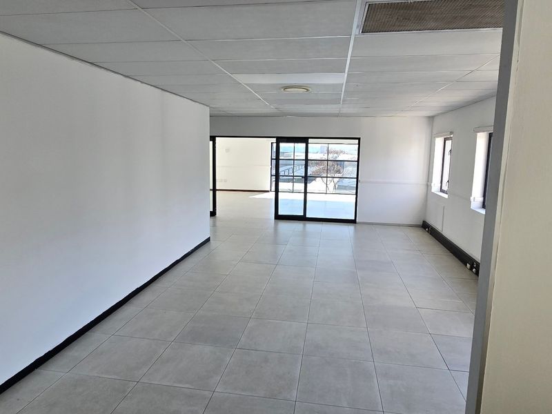 A bright, vacant commercial office space featuring neutral tiled flooring, a suspended ceiling, and sliding glass doors leading to an outdoor area.