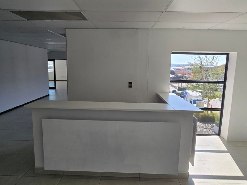 An open-plan office space featuring a built-in reception desk, tiled flooring, and large windows providing natural light.