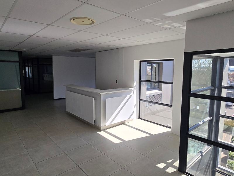 Open-plan commercial office space featuring a built-in reception desk, tiled flooring, suspended ceiling, and large windows with natural light.