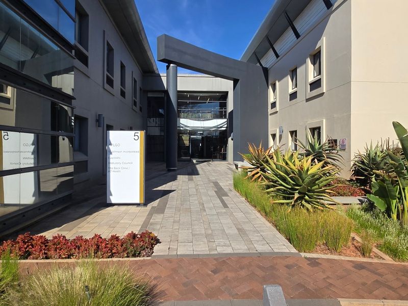 Modern commercial building entrance featuring a paved walkway, glass facade, and well-maintained landscaped garden beds.