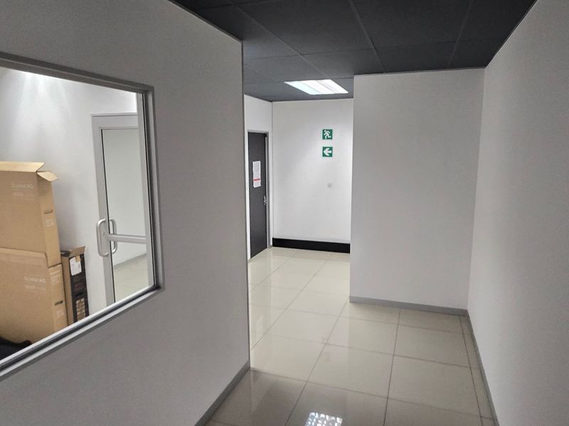 A clean, modern commercial hallway featuring tiled flooring, a suspended ceiling with recessed lighting, and an internal glass partition looking into an adjacent office space.