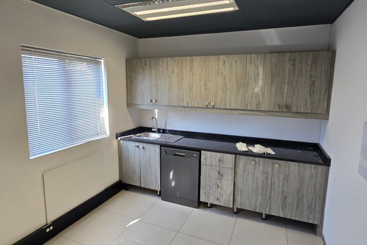 A clean, functional kitchenette featuring wood-look cabinetry, dark countertops, a stainless steel sink, and an integrated dishwasher in a bright, tiled office space.