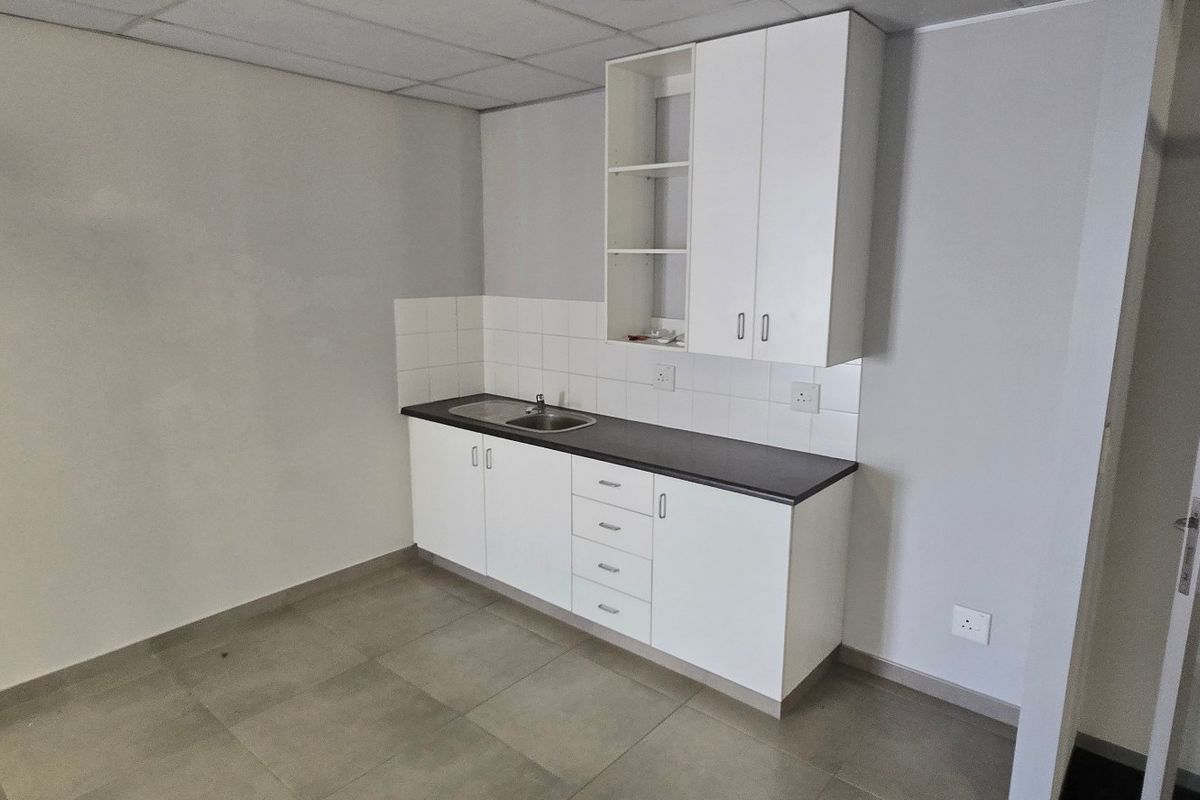 A clean, compact kitchenette featuring white built-in cabinetry, a dark laminate countertop, stainless steel sink, and neutral tiled flooring in a commercial space.