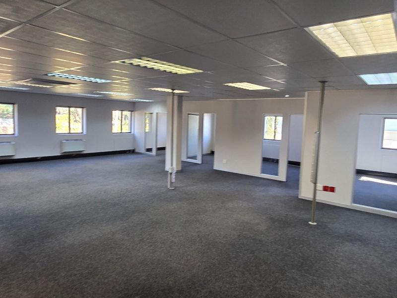 A spacious, open-plan commercial office area featuring a suspended ceiling, recessed lighting, carpeted flooring, and wall-mounted HVAC units.
