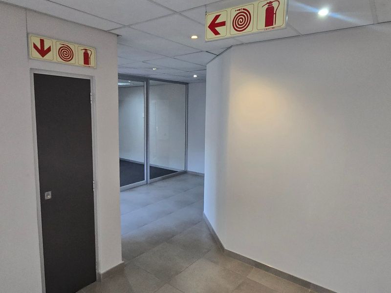 A clean, modern commercial hallway featuring tiled flooring, a suspended ceiling with recessed lighting, and glass partition walls leading to office space.