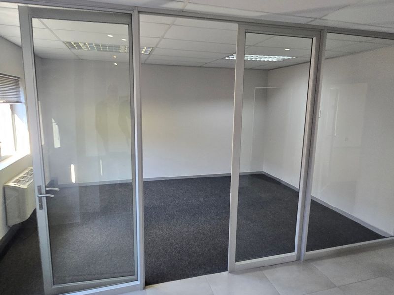 A clean, vacant commercial office space featuring modern glass partitioning, suspended ceilings, recessed lighting, and carpeted flooring.