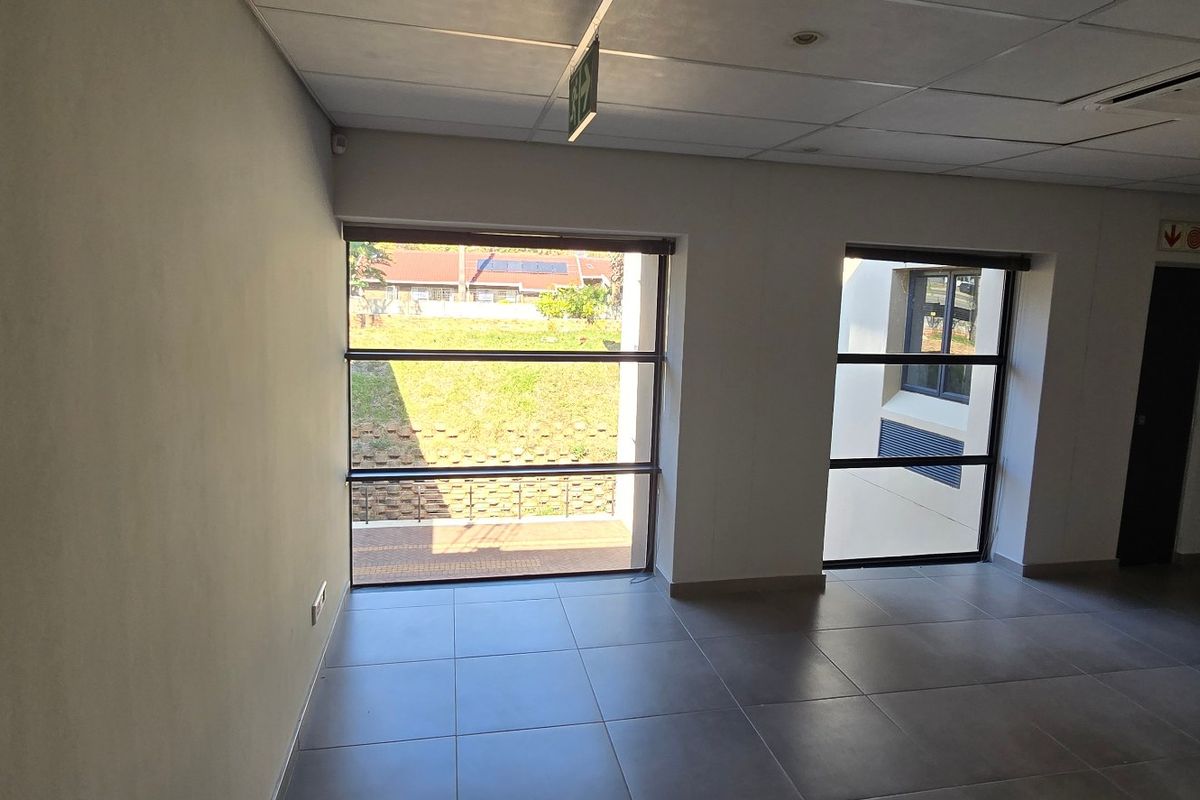 A bright, empty commercial office space featuring tiled flooring, a suspended ceiling with recessed lighting, and large windows providing natural light.