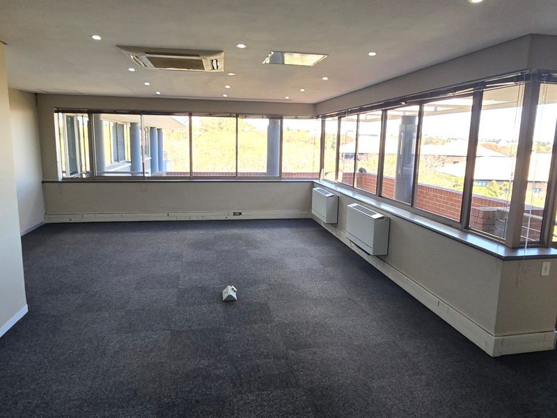 A spacious, empty commercial office area featuring large wrap-around windows, carpeted flooring, recessed lighting, and integrated ceiling and wall-mounted air conditioning units.