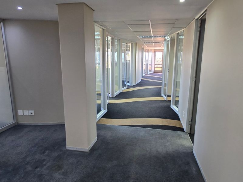 A bright, modern commercial office hallway featuring glass partition walls, recessed lighting, and patterned carpet flooring, creating a professional and open workspace environment.