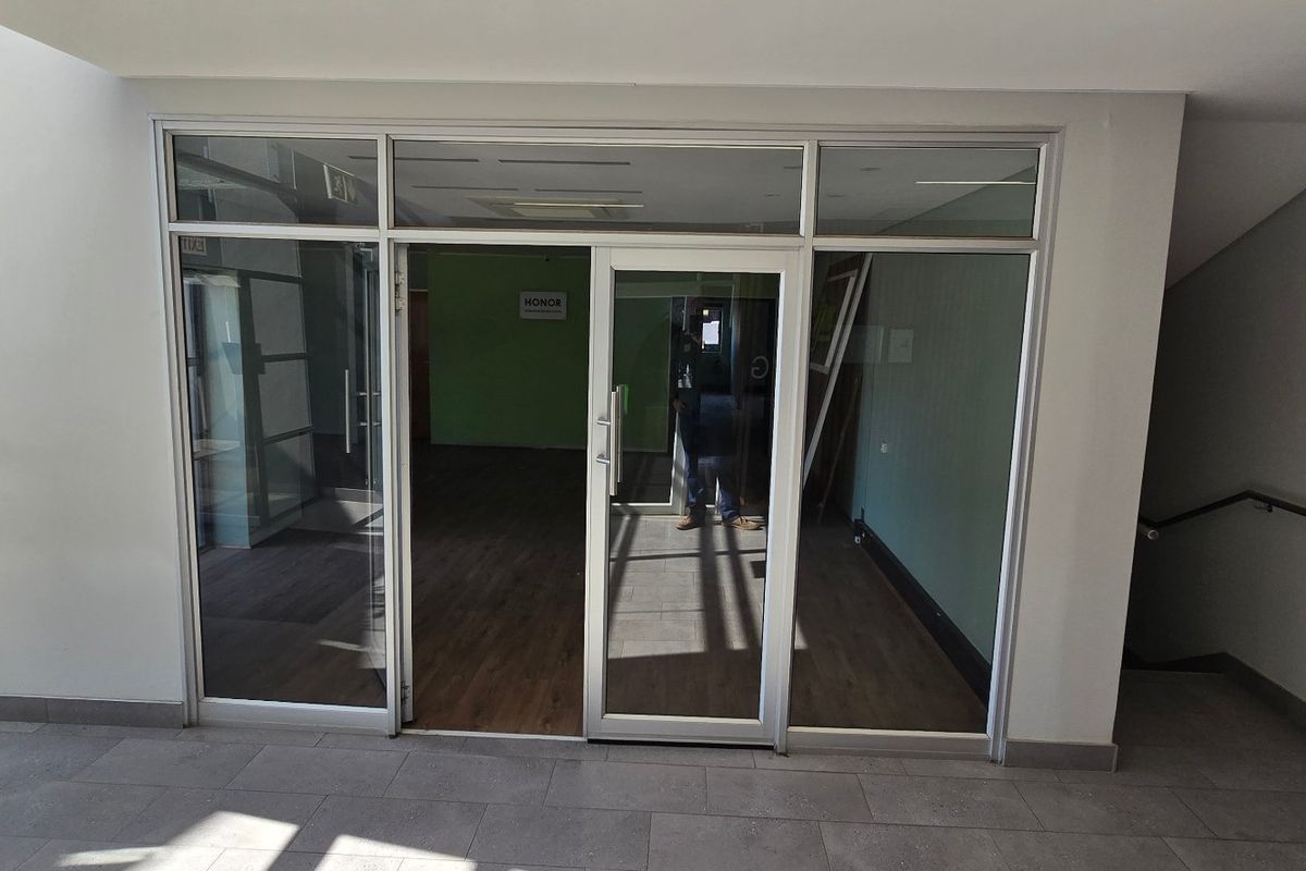 Modern commercial office entrance featuring aluminium framed glass doors and partitioning, leading into an open-plan workspace with wood-look flooring.