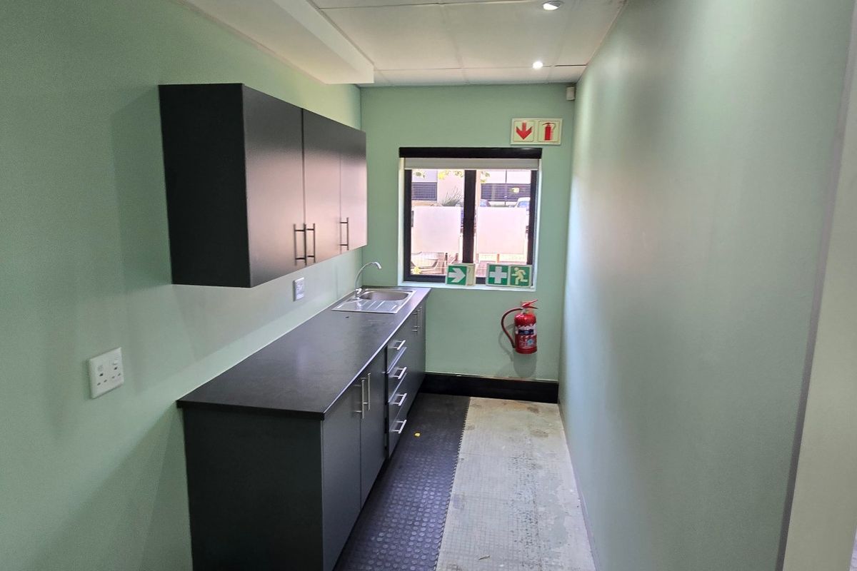 A compact kitchenette area featuring dark cabinetry, a stainless steel sink, and functional emergency safety equipment in a commercial office space.