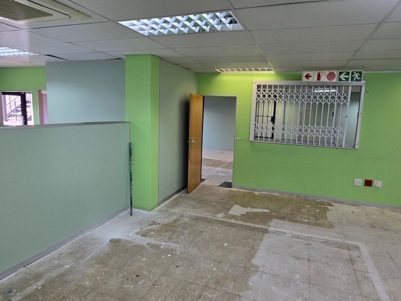 An empty commercial office space featuring a suspended ceiling, recessed lighting, security gate, and partition walls, requiring flooring and wall finish updates.