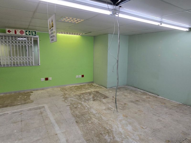 An empty commercial office space featuring a suspended ceiling, fluorescent lighting, and a security gate, currently requiring floor and wall surface repairs.