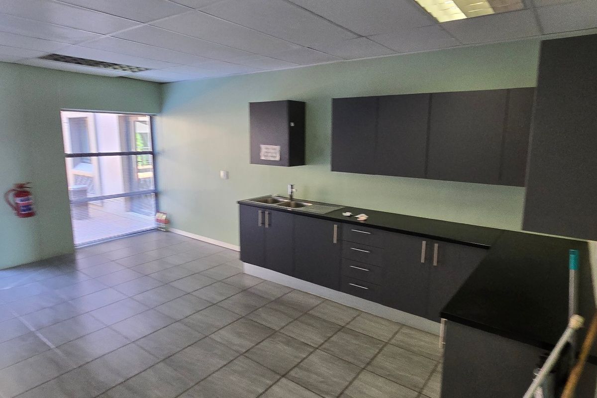 A clean, functional kitchenette area featuring modern dark cabinetry, a stainless steel sink, tiled flooring, and ample natural light from large windows.