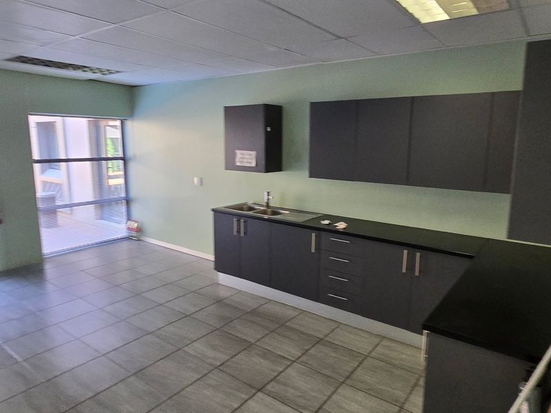A clean, functional kitchenette area featuring modern dark cabinetry, a stainless steel sink, tiled flooring, and ample natural light from large windows.