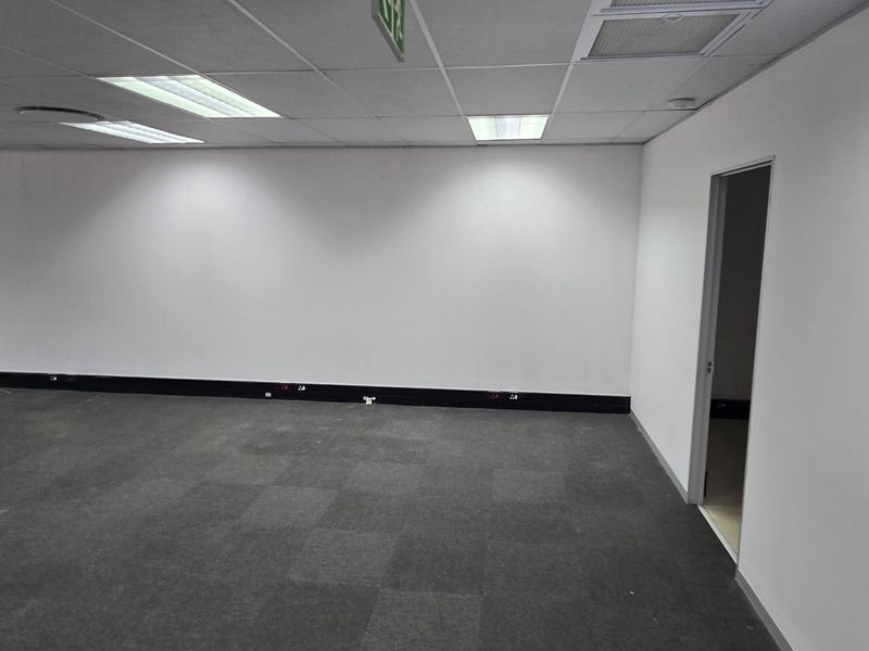 A vacant, open-plan commercial office space featuring neutral walls, carpet tile flooring, and a suspended ceiling with recessed lighting.