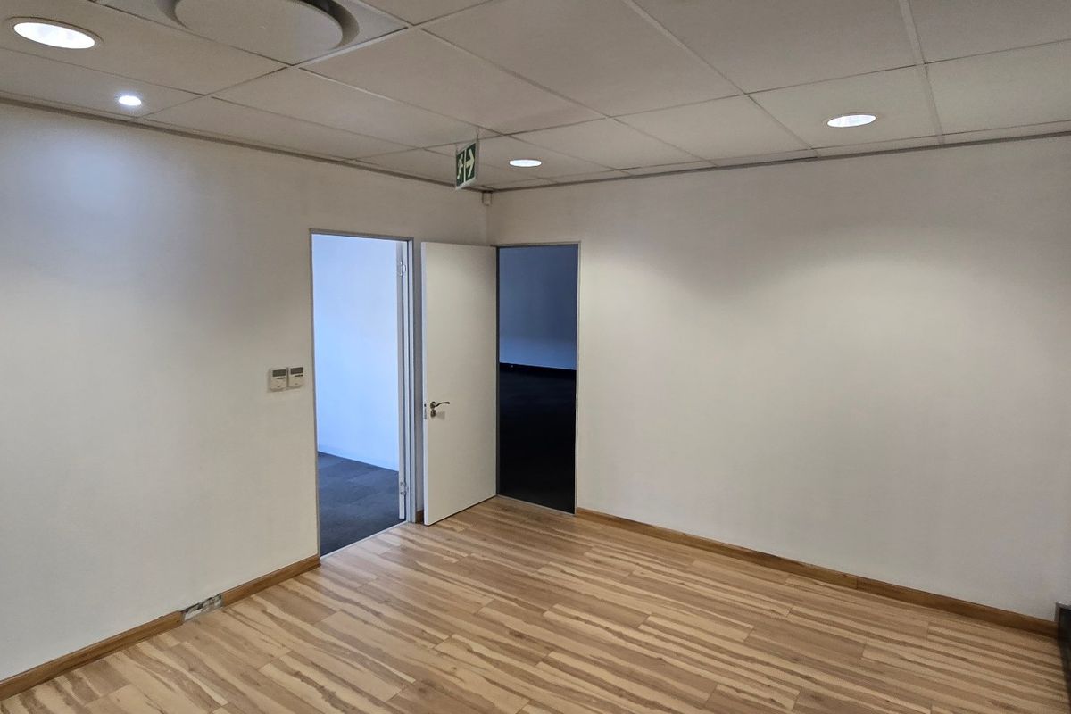 A vacant, well-lit office space featuring laminate flooring, a suspended ceiling with recessed lighting, and two doorways leading to adjacent areas.