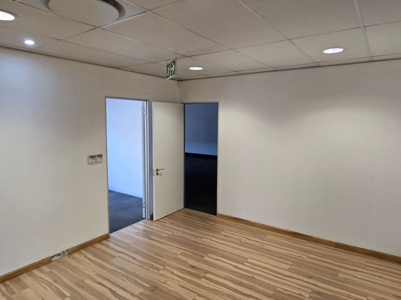 A vacant, well-lit office space featuring laminate flooring, a suspended ceiling with recessed lighting, and two doorways leading to adjacent areas.