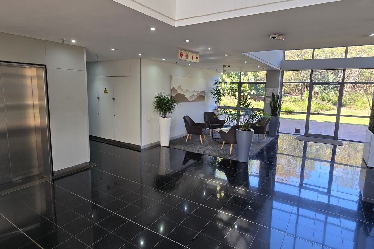 Modern commercial building entrance lobby featuring polished dark tile flooring, a stainless steel elevator, and expansive glass windows overlooking the surrounding greenery.