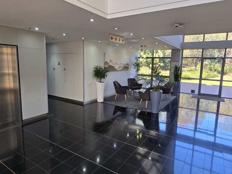 Modern commercial building entrance lobby featuring polished dark tile flooring, a stainless steel elevator, and expansive glass windows overlooking the surrounding greenery.