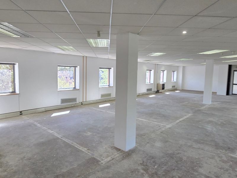 Spacious open-plan commercial office area featuring a suspended ceiling, recessed lighting, structural columns, and a small kitchenette area with a sink.