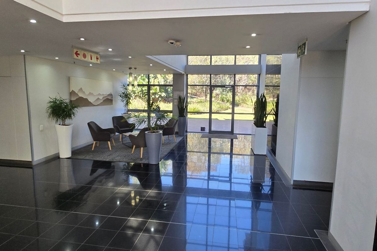 Spacious commercial building entrance lobby featuring polished dark floor tiles, recessed lighting, and floor-to-ceiling glass panels providing natural light and garden views.