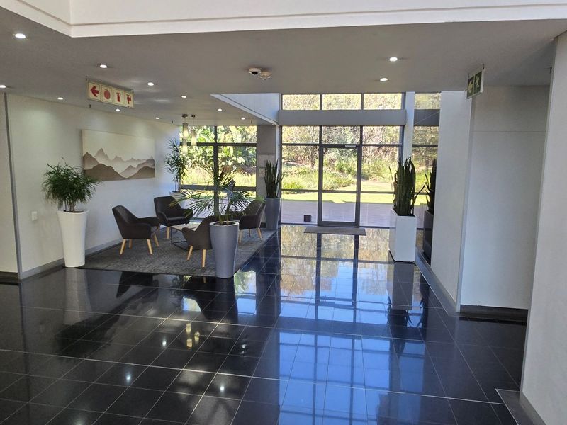 Spacious commercial building entrance lobby featuring polished dark floor tiles, recessed lighting, and floor-to-ceiling glass panels providing natural light and garden views.