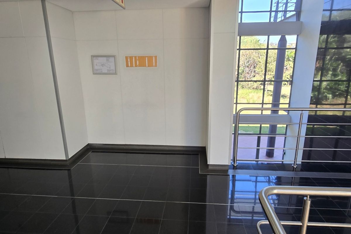A modern commercial hallway featuring polished black floor tiles, a stainless steel balustrade, and a large glass facade providing natural light.