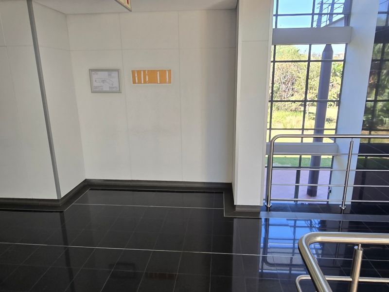 A modern commercial hallway featuring polished black floor tiles, a stainless steel balustrade, and a large glass facade providing natural light.