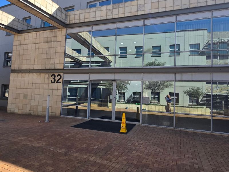 Modern commercial building entrance featuring a glass curtain wall facade, stone cladding, and a paved forecourt area.