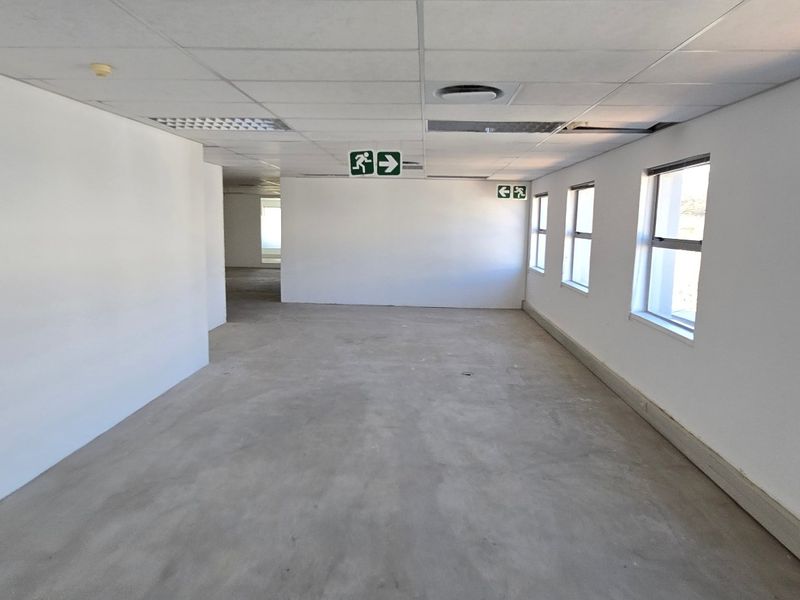 An empty, open-plan commercial office space featuring a suspended ceiling, fluorescent lighting, and large windows, currently requiring flooring and ceiling maintenance.