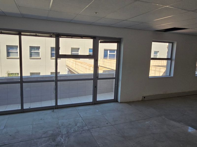 An empty commercial office space featuring tiled flooring, a suspended ceiling, and large windows providing natural light and access to an exterior balcony.