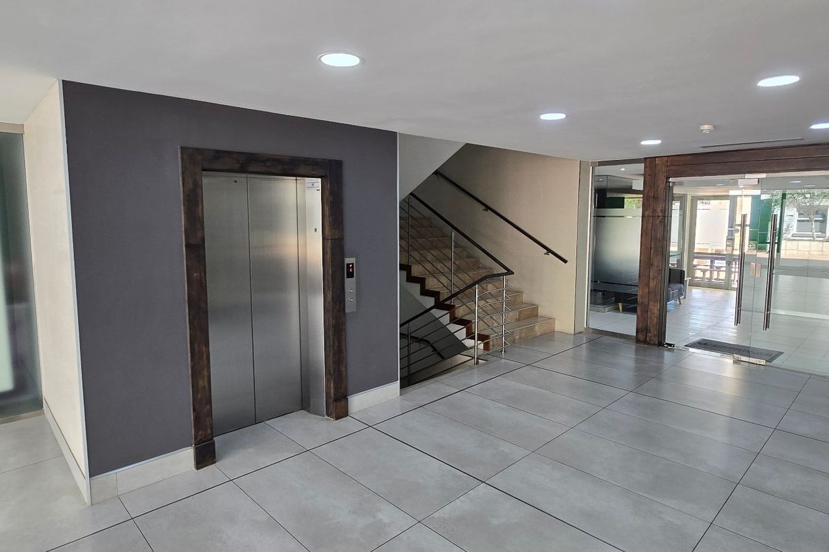 Modern commercial building entrance lobby featuring tiled flooring, a stainless steel elevator, and a staircase with a contemporary handrail.