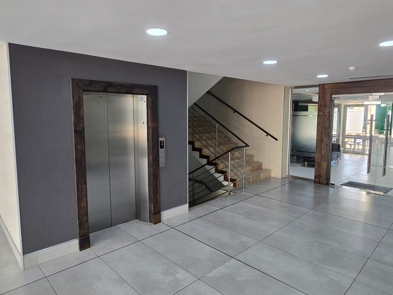 Modern commercial building entrance lobby featuring tiled flooring, a stainless steel elevator, and a staircase with a contemporary handrail.
