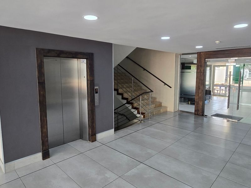Modern commercial building lobby featuring a stainless steel elevator, staircase, and glass entrance doors with tiled flooring and recessed lighting.