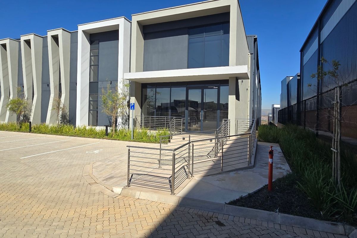 Modern industrial building exterior featuring a glass facade, paved parking area, and accessible metal ramp with stainless steel railings.