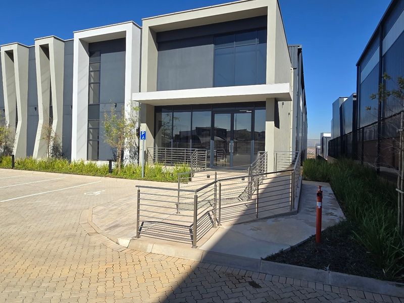 Modern industrial building exterior featuring a glass facade, paved parking area, and accessible metal ramp with stainless steel railings.